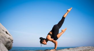 Woman performing a yoga pose on a rocky beach with ocean view and clear blue sky.